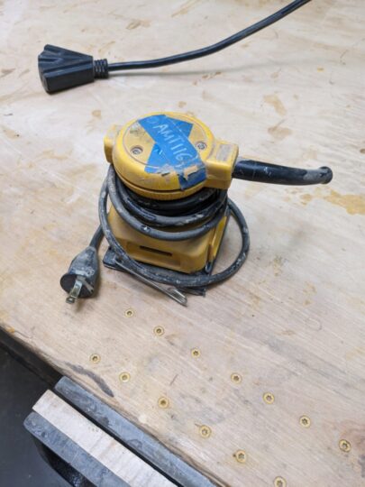 The yellow and black palm sander wrapped in power cable on top of work bench