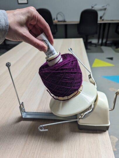 A folded yarn label placed in the divot of the cone of a yarn winder filled with a purple cake of yarn.