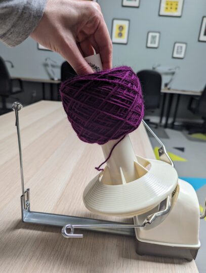 A purple cake of yarn being transferred from the cone of the ball winder onto the folded label that originally came with the yarn.