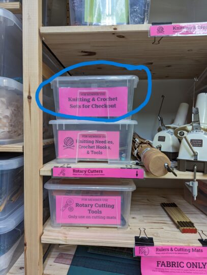 A close-up photo of the Knitting and Crochet Sets for Checkout bin on the textiles shelving. It is circled in blue and located to the left of the yarn swift and ball winder.