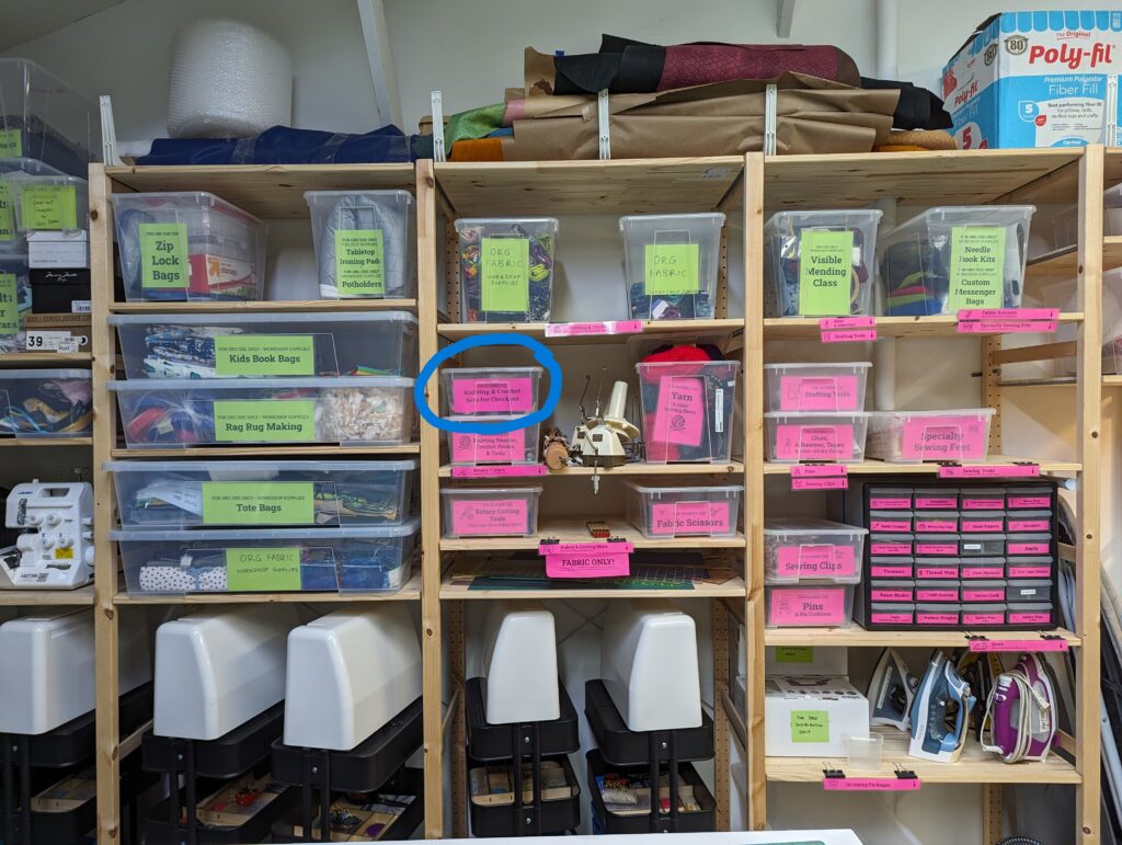 Photo of The Knitting and Crochet Sets for Checkout bin, circled in blue, on the textiles walls. 